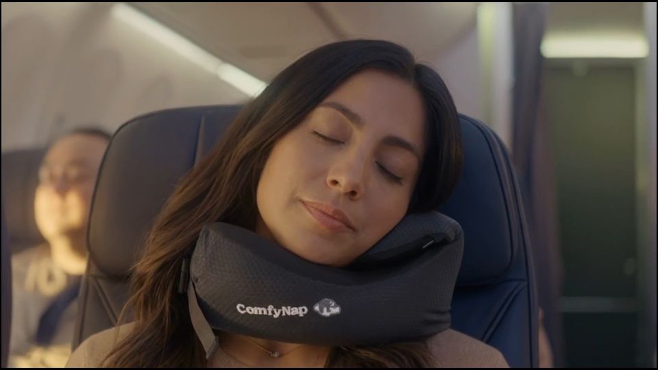 A passenger asleep in her airplane seat, her head held by the black ComfyNap pillow
